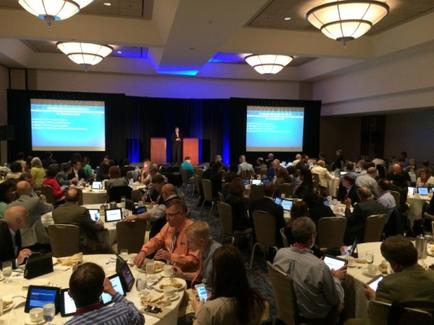 A large conference room with a high ceiling and chandeliers. The room is filled with people sitting at long tables with laptops in front of them. The tables are covered with white tablecloths and there are plates cups and saucers on the tables. In the center of the room there is a stage with a podium and a blue curtain. On either side of the stage there are two large screens displaying text. The people in the room appear to be engaged in a discussion or presentation.