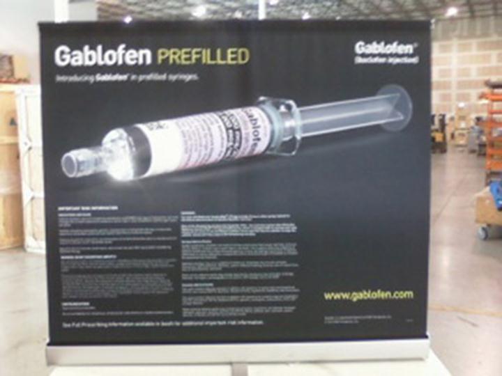 Of a black banner with the text "Gablofen Prefilled" and an image of a syringe. The banner is displayed in a warehouse or storage area with shelves and boxes in the background. The syringe is shown in the center of the banner with the needle pointing towards the right side of the image. The text on the banner is in white and black and it appears to be an advertisement for the product. The website "www.gablofen.com" is also visible on the bottom right corner.
