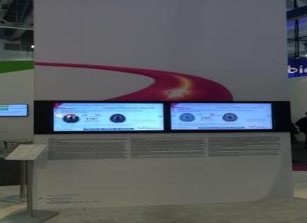 A large white display stand with two large screens on it. The screens are displaying information about a website or app. The background of the display stand is white with a red curved design on the top left corner. On the right side of the screen there is a smaller screen with a blue and white image of a person's face. Below the screens there are several smaller screens with information about the website's features. The stand is located in an exhibition hall with other exhibits visible in the background.