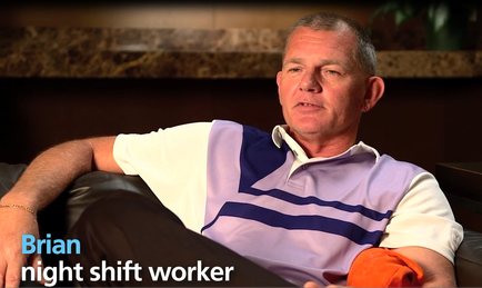 A still from a video interview with Brian Night Shift Worker. He is sitting on a couch with his legs crossed and his arms resting on the armrests of the couch. Brian is wearing a purple and white striped polo shirt and black pants. He has short grey hair and is looking directly at the camera with a serious expression on his face. The background is blurred but it appears to be a living room with a fireplace and a plant. The text on the image reads "Brian night shift worker".