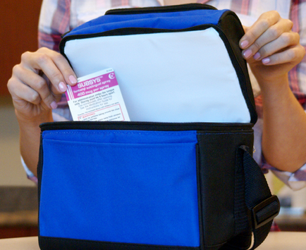 A person holding a blue and black lunch bag with a white label on it. The bag appears to be made of a durable material and has a handle on the top for easy carrying. The person is holding the bag with both hands and the label is visible on the front of the bag. The background is blurred but it seems to be an indoor setting with a wooden table and chairs visible.