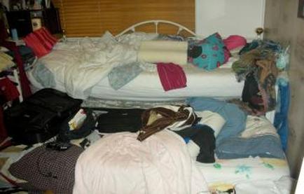 A messy bedroom with a white bed in the center. The bed is covered in a white comforter and there are several pillows and blankets scattered around it. On the left side of the bed there is a pile of clothes bags and other miscellaneous items. The room appears to be cluttered and disorganized. The walls are painted white and the floor is made of wood. There is a window with blinds in the background.