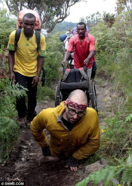 A group of three men hiking on a trail in a forested area. The man in the foreground is wearing a yellow jacket and a red bandana on his head and he is crouching down on the ground with his hands on his knees. He appears to be searching for something. Behind him there is another man wearing a red t-shirt and carrying a large backpack on his back. He is pushing a wheelchair with a backpack attached to it. The other two men are walking behind him also carrying backpacks. The trail is covered in greenery with trees and bushes on either side. The sky is overcast and the overall mood of the image is somber.