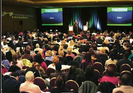A large conference room with a stage in the center. The room is filled with people sitting in rows of chairs facing the stage. On the stage there is a man standing at a podium and speaking into a microphone. Behind him there are two large screens displaying the text "Q&A" and "Exchange". The room appears to be well-lit with natural light coming in from the windows on the left side of the image. The people in the room are attentively listening to the speaker.