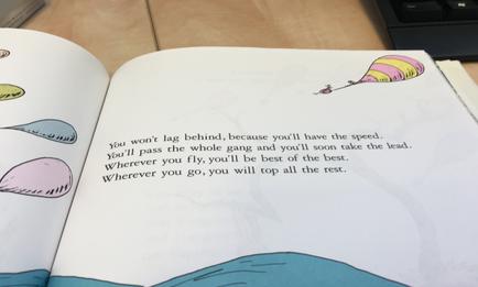 An open book with a page from a children book. The page is divided into two sections. On the left side of the page there is a cartoon illustration of a pink and yellow hot air balloon with a quote written on it that reads "You won't lag behind because you'll have the speed. You'll pass the whole gang and you'll soon take the lead. Wherever you fly you'll be best of the best. wherever you go you will top all the rest." The background is a light blue sky with white clouds. The book is resting on a wooden desk with a keyboard visible in the background.