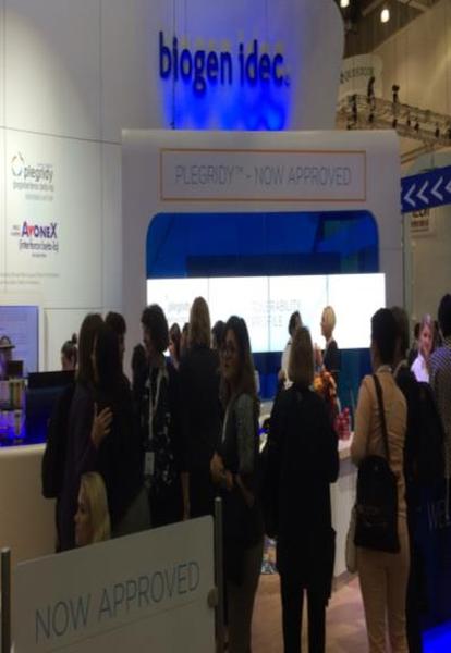 A group of people standing in front of a booth at a trade show. The booth has a blue and white color scheme with the word "biogen idea" written in bold letters at the top. Below the word there is a sign that reads "Plegrady - Now Approved" in white letters. The people in the image appear to be engaged in conversation and are looking at the booth. There is a blue arrow pointing to the right side of the booth indicating that the booth is now approved. In the background there are other booths and people walking around.