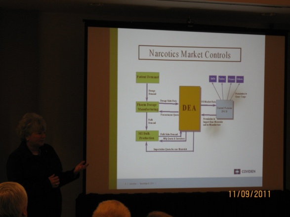 A woman giving a presentation next to a projection screen. The image on the screen is a flow chart with the title Narcotics Market Controls. An orange box in the middle represents the DEA (Drug Enforcement Administration) with 3 green boxes to the left and 1 blue box & 4 purple boxes to the right all connected by arrows & supplemented by annotations. The logo of Covidien is in the bottom right corner along with a date on the photograph-proper (11/09/2011).