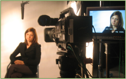 A woman sitting in front of a large video camera. She is wearing a black blazer and appears to be in a studio or recording studio. The camera is mounted on a tripod and is positioned in the center of the image. On the right side of the camera there is a large screen with a picture of a woman's face on it. The background is a white wall with a light shining on the camera. The woman is looking directly at the camera with a serious expression on her face.