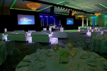 A large event space with a high ceiling and chandeliers hanging from it. The room is set up for a formal event with white tables and chairs arranged in a semi-circle. The tables are covered with white tablecloths and have gold chargers plates and cutlery on them. In the center of the room there is a stage with a large screen that reads "Better Together" and a podium with a microphone. The stage is decorated with blue and green lights creating a warm and inviting atmosphere. The overall color scheme of the space is blue green and white.