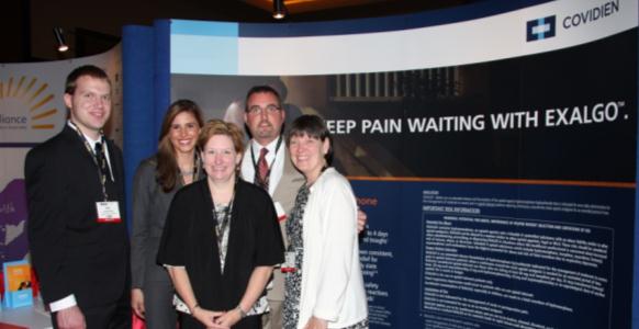 A group of five people standing in front of a blue and white banner with the text "Keep pain waiting with Exalgo" and the logo of Covidien. The banner is located at an exhibition or trade show booth. The people in the photo are smiling and appear to be posing for a photo. The background is dark and there are other banners visible in the background.