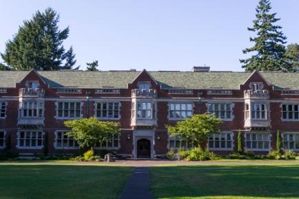 A large two-story brick building with a green roof. The building has multiple windows and balconies on the second floor with a large entrance in the center. The entrance is flanked by two columns on either side. There are trees and shrubs surrounding the building and a grassy lawn in front of it. The sky is blue and there are a few clouds in the distance. A walkway leads up to the entrance of the building.