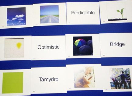 A collage of nine square cards arranged in a grid-like pattern on a blue background. Each card has a different image on it including a photograph of a road a blue sky with white clouds a green plant and a rainbow-colored umbrella. The first card on the top left shows a photograph with a yellow sun in the sky. The second card has the word "Optimistic" written in white text. The third card has an image of a person holding a dog the fourth card has two people holding hands and the fifth card has three people walking in the background. The sixth card has text that reads "Predictable" and "Bridge". The seventh card has another image of the same person holding an umbrella the eighth card has images of a group of people walking together the ninth card has pictures of a dog and a person playing with a ball the tenth card has color swatches of different colors the eleventh card has green squares the twelfth card has yellow squares and all the cards have text written on them.