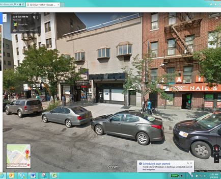 A street view of a building on a corner of a street. The building appears to be a two-story brick building with a flat roof and multiple windows. There are several shops and restaurants on the corner of the street including a nail salon and a nail shop. The street is lined with trees and there are several cars parked on the side of the road. The sky is blue and the street is empty with no people visible. The image is taken from a top-down perspective looking down on the street.