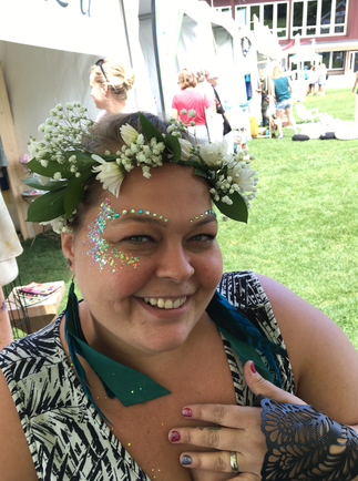 A close-up portrait of a young woman with a flower crown on her head. She is smiling and looking directly at the camera. The woman is wearing a black and white patterned dress with a blue ribbon tied around her neck. She has a tattoo on her left arm and her right hand is resting on her shoulder. The flower crown is made up of white flowers and green leaves. The background appears to be an outdoor event with tables and chairs set up and people in the background. The overall mood of the image is happy and relaxed.