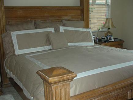 A bedroom with a large bed in the center. The bed has a wooden headboard and footboard and is covered with a beige comforter and several beige pillows. On the right side of the bed there is a nightstand with a lamp a phone and a picture frame. The nightstand is made of wood and has a carved design on the top. The room has a window with white blinds and the walls are painted a light beige color.