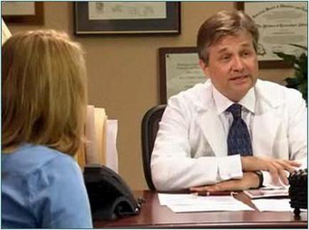 A man and a woman sitting at a desk in an office. The man is wearing a white lab coat and a blue tie and he appears to be a doctor or a medical professional. He is sitting in front of a desk with papers and a telephone on it and there is a framed certificate on the wall behind him. The woman is sitting next to him and she is looking at him attentively. It seems like they are engaged in a conversation.