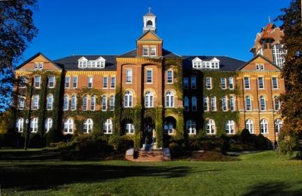 A large two-story brick building with a clock tower on top. The building has multiple windows and is covered in ivy. The sky is blue and there are trees on either side of the building. In front of the clock tower there is a small fountain with a statue of a man on it. The ground is covered with grass and there is another building visible in the background. The overall atmosphere of the image is peaceful and serene.