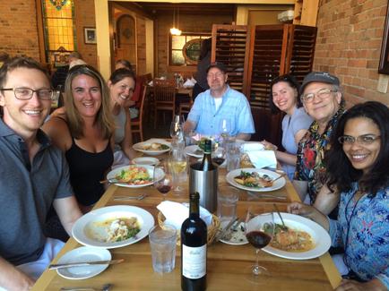 A group of nine people sitting at a dining table in a restaurant. They are all smiling and appear to be enjoying their meal. The table is set with plates of food glasses of wine and a bottle of wine. The restaurant has brick walls and wooden tables and there is a large window in the background. The people are dressed in casual clothing and some are wearing hats. The overall atmosphere of the restaurant is relaxed and casual.