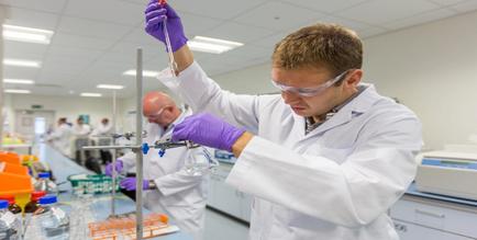 A young man in a laboratory setting. He is wearing a white lab coat goggles and purple gloves and is holding a pipette in his right hand and a test tube in his left hand. He appears to be conducting a scientific experiment. In front of him there are several other people working in the background also wearing lab coats and gloves. On the left side of the image there is a long table with various laboratory equipment and supplies. The man is holding the pipette with both hands and is looking down at it intently. He seems to be focused on his work. The background is blurred but it seems like the laboratory is well-lit with fluorescent lights.