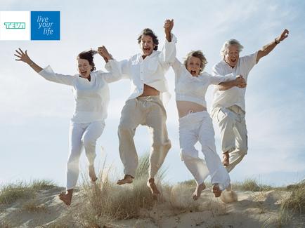 Four people two men and two women jumping in the air with their arms stretched out wide. They are all wearing white clothes and appear to be in a desert-like environment with sand dunes and grass in the background. The sky is blue and the sun is shining suggesting that it is a sunny day. The people are smiling and seem to be having a great time. The image also has a logo that reads "Live your life" in the top left corner.