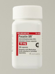 A bottle of 100 Focalin XR (dexmethylphenidate HCl) extended-release capsules CII in the 10 mg dosage manufactured by Novartis. The bottle cap and label are all white. The label has graphic elements in red and black with text in black and white. The label instructs pharmacists to "Dispense with Medication Guide attached or provided separately". The background is plain gray.