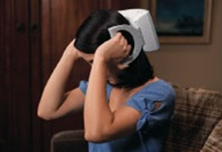 A young woman sitting on a couch with her head in her hands. She is wearing a blue t-shirt and appears to be upset or frustrated. On top of her head there is a white hair dryer that is covering her eyes. The background is blurred but it seems like the woman is in a living room with a wooden cabinet and a painting hanging on the wall.