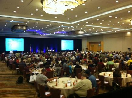 A large conference room with a high ceiling and a chandelier hanging from it. The room is filled with people sitting at round tables facing a stage where a man is giving a presentation. The stage is covered with a blue curtain and there are two large screens on either side of the stage. The people in the room appear to be engaged in the presentation with some sitting at the tables and others standing at the front of the room. The overall atmosphere is formal and professional.