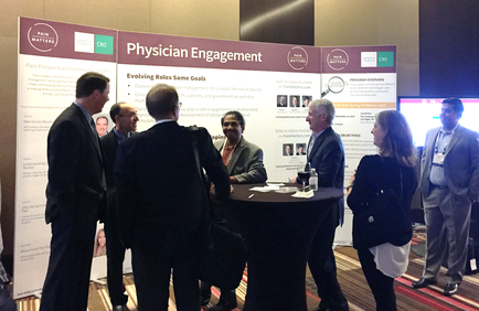 A group of people gathered around a table in front of a large banner that reads "Physician Engagement". The banner also has images and text on it likely related to the topic of physician engagement. There are six people in the image six men and one woman all dressed in formal business attire. They appear to be engaged in a conversation with one man gesturing with his hands as he speaks. The background is a conference room with a large screen on the right side of the image.