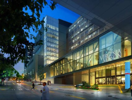 A digital rendering of a modern building at night. The building is located on a street corner with a busy intersection in front of it. The street is lined with trees and there are people walking on the sidewalk. The sky is dark and the building is lit up with bright lights. On the right side of the image there is a large glass facade with the words "Virginia Mason" written in white letters. The entrance of the building has a sign that reads "Welcome to Virginia Mason." The building appears to be a high-rise with multiple floors and large windows.