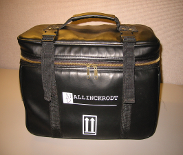 A black leather doctor bag with Mallinckrodt branding on its side. It is rectangular in shape with a double brass zipper (allowing the lid to flip open) two vertical straps (providing additional support) and a handle on top (for carrying in a single hand). This was a promotional item presumably. It was photographed on a flat surface against a beige background.