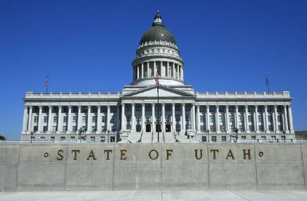 Of the Utah State Capitol building in Salt Lake City Utah. The building is a large neoclassical structure with a dome on top and columns on either side. The dome is topped with a statue of a man holding a flag. In front of the building there is a concrete wall with the words "State of Utah" inscribed on it. The sky is blue and there are trees in the background. The image appears to have been taken on a sunny day.