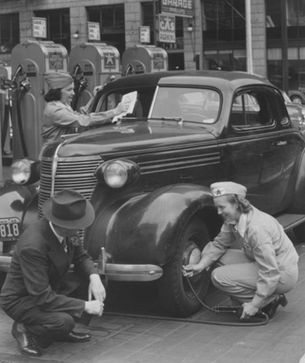 A black and white photograph of three people at a gas station. The car is a vintage model with a large front grille and a curved roof. It is parked on the side of the street and there are several gas pumps visible in the background. In the foreground there are two men and a woman working on the car. The man on the left is wearing a suit and a hat and the woman on the right is holding a clipboard and appears to be filling up the car with fuel. The woman is standing next to the car and she is looking at the man with a smile on her face. The two men are kneeling on the ground and one of them is using a hose to clean the car's tires. The background shows a building with a sign that reads "Gas Station."