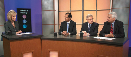 Four people sitting at a desk in a news studio. They are all dressed in formal business attire and appear to be engaged in a conversation. On the left side of the desk there is a woman with blonde hair wearing a black blazer and a white blouse. She is sitting at the desk with a microphone in front of her. Next to her there are two men one of whom is wearing a suit and tie and the other two are wearing suits and ties. The man in the middle is holding a pen and appears to be speaking while the man on the right is listening attentively. The desk is covered with papers and there are coffee mugs on it. The background of the image is a blue wall with a large screen displaying the text "The Role of History".