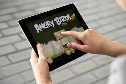 A person holding an iPad in their hands. The iPad screen displays the game "Angry Birds Free" with the title of the game in bold yellow letters at the top. Below the title there is a green background with a cartoon-like illustration of a bird flying over a grassy field. On the right side of the screen there are three buttons - a yellow button a green button and a red button. The person's hand is pointing towards the screen with their index finger. The background is blurred but it appears to be a brick floor.