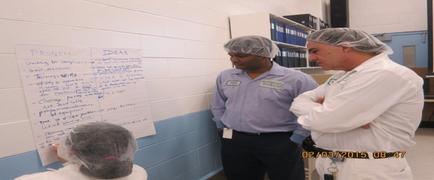 Three men in a laboratory setting. They are all wearing white lab coats and hairnets on their heads. The man in the middle is wearing a blue shirt and is standing with his arms crossed while the man on the right is standing next to him. On the left side of the image there is a whiteboard with a list of tasks written on it. The tasks appear to be related to problem-solving and ideas. The date "02/03/2015 08:47" is visible in the bottom right corner.