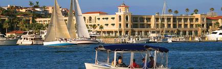 A marina with several boats and yachts docked in the water. In the foreground there is a white boat with a blue canopy and a group of people on board. The boat is sailing on the water and there are several other boats in the background. On the left side of the image there are two sailboats with white sails and on the right side there appears to be a large building with a red roof and a clock tower. The building is surrounded by palm trees and other buildings. The sky is blue and the water is calm and the overall scene is peaceful and serene.
