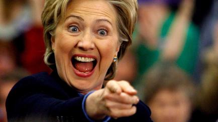 A close-up portrait of former US Secretary of State Hillary Clinton. She is pointing her finger towards the camera with a surprised or shocked expression on her face. Her eyes are wide open and her mouth is slightly open as if she is shouting or making a point. She has blonde hair and is wearing a dark blue blazer. The background is blurred but it appears to be a crowd of people.