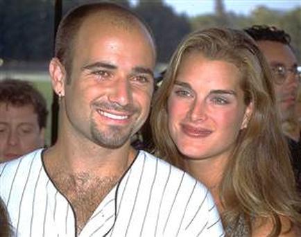 A man and a woman posing for a photo together. The man is on the left side of the image and the woman on the right side. They are both smiling and looking at the camera. The woman has long blonde hair and is wearing a black dress with a plunging neckline. She has a tattoo on her chest and is also smiling. The background appears to be a crowd of people with some people visible in the background. It seems like they are at an event or gathering.
