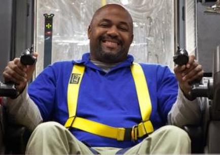 A man sitting in a chair and holding two guns in his hands. He is wearing a blue polo shirt and yellow safety harness. He has a big smile on his face and appears to be happy and relaxed. The background is blurred but it seems like he is inside a room with a window and some equipment. The man is holding the guns with both hands and is looking directly at the camera.