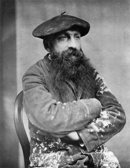 A black and white portrait of a man with a long beard and mustache. He is sitting on a chair with his arms crossed in front of him. He has a serious expression on his face and is looking directly at the camera. The man is wearing a flat cap with a feather on top and a striped shirt underneath. The background is plain and the overall mood of the image is somber.