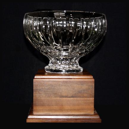 Of a glass bowl on a wooden base. The bowl is round and has a wide base with a narrow neck and a wide opening at the top. It appears to be made of clear glass with a wavy pattern on the sides. The base is square in shape and is made of a dark wood with a smooth finish. The background is black making the bowl stand out.
