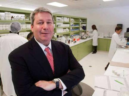 A man in a suit and tie standing in a laboratory. He is standing with his arms crossed and is smiling at the camera. He appears to be in his late 40s or early 50s. Behind him there are shelves stocked with various laboratory equipment and supplies. On the right side of the image there is a desk with a computer monitor and other scientific equipment. In the background two people wearing white lab coats can be seen working in the laboratory. The overall atmosphere of the laboratory is professional and organized.