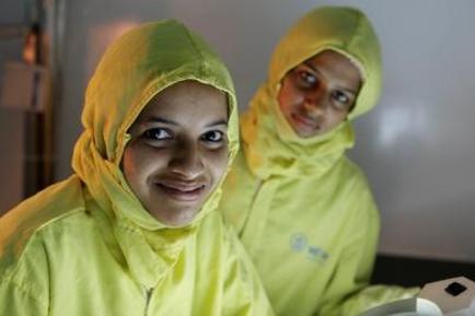 Two young women wearing bright yellow raincoats. They are standing in a room with a white wall and a door in the background. The woman on the left is smiling and looking directly at the camera. She is holding a white object in her hand which appears to be a remote control or a device. The other woman is also smiling and appears to have a serious expression on her face. Both women are wearing the same yellow raincoat but their hair is pulled back in a neat bun.