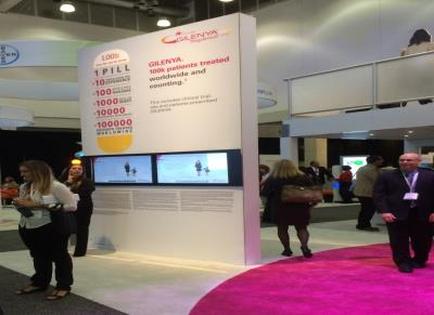 A trade show booth with a pink carpeted floor and white walls. The booth has a large screen on the right side displaying information about the company's products and services. On the left side of the booth there is a sign that reads "100% PILL" and below it there are three smaller screens displaying the company name and contact information. There are several people in the background some of whom are looking at the booth and others are engaged in conversation. The overall atmosphere of the image is busy and bustling.