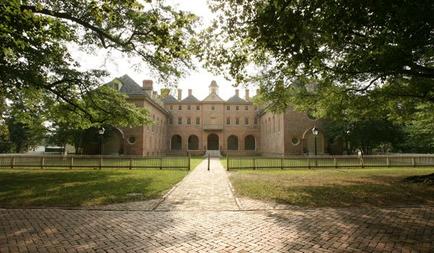 A large brick building with multiple levels and arched windows. The building appears to be a college or university campus as there are trees and greenery surrounding it. In front of the building there is a brick pathway leading up to the entrance. The sky is blue and the sun is shining through the trees casting shadows on the ground. The overall atmosphere of the image is peaceful and serene.