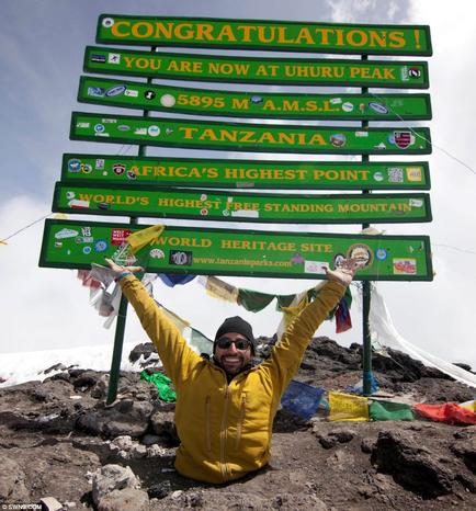 A man sitting on top of a mountain with his arms raised in the air. He is wearing a yellow jacket sunglasses and a beanie. Behind him there is a large green signpost with multiple signs pointing in different directions. The signs read "Congratulations! You are now at Uhuru Peak Tanzania Africa's highest point World's highest standing mountain World Heritage Site". The signpost is located on a rocky terrain with a clear blue sky in the background. There are also colorful flags and banners hanging from the poles. The man appears to be happy and proud of his accomplishment.