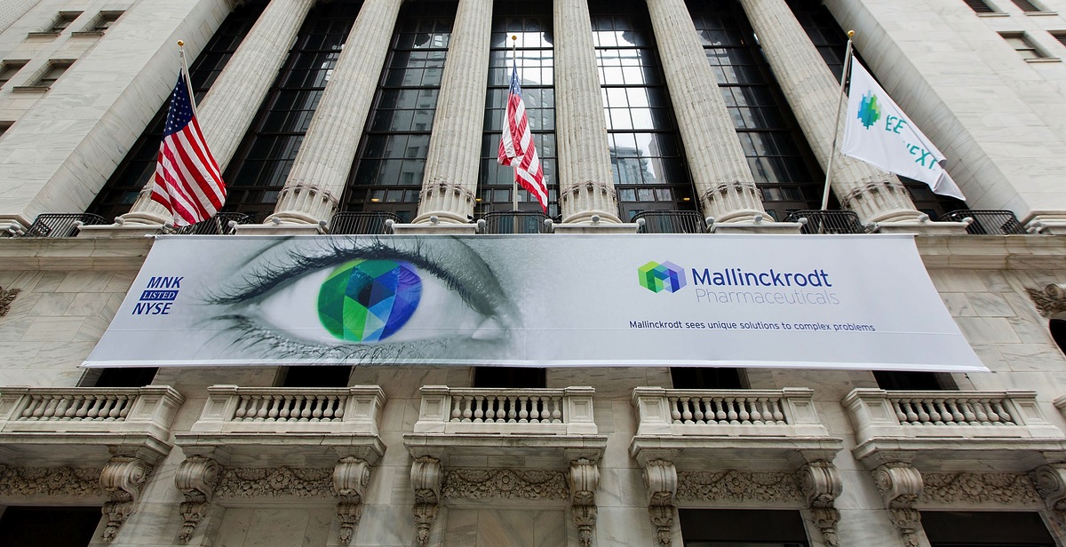 The exterior of the New York Stock Exchange Building (a.k.a. the NYSE Building) located in the Financial District of Lower Manhattan in New York City. There are 3 flags (2 American flags and 1 unidentified flag) and a large banner for Mallinckrodt Pharmaceuticals (with the tagline "Mallinckrodt sees unique solutions to complex problems"). The banner also includes the stock ticker symbol for Mallinckrodt Pharmaceuticals (MNK) a computer drawing of an eye in which the iris is rendered to resemble the logo of Mallinckrodt Pharmaceuticals and the logo of Mallinckrodt Pharmaceuticals.
