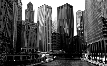 A black and white photograph of a city skyline. The photo is taken from a low angle looking down on a river with a bridge crossing over it. The river is lined with tall buildings on both sides and there are trees on the left side of the image. In the center of the photo there is a tall skyscraper with a dome on top. The building appears to be made of glass and steel and it is surrounded by other skyscrapers. The sky is overcast and the overall mood of the photograph is gloomy and gloomy.