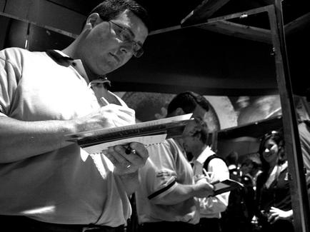 A black and white photograph of a man standing in a room with other people in the background. The man is wearing a polo shirt and glasses and is holding a clipboard in his hands. He appears to be writing or drawing on the clipboard with a pen. He is looking down at the clipboard and seems to be focused on his work. The room has a high ceiling and there is a metal railing on the right side of the image. The people in front of him are also writing or taking notes.