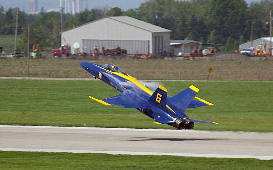A blue and yellow fighter jet taking off from an airport runway. The jet is in the center of the image with its wings spread wide and its nose pointed upwards. The tail of the jet is painted in the colors of the United States Navy with the number 6 on the side. In the background there is a large hangar and a construction site with various vehicles and equipment. The sky is overcast and there are trees and buildings visible in the distance.
